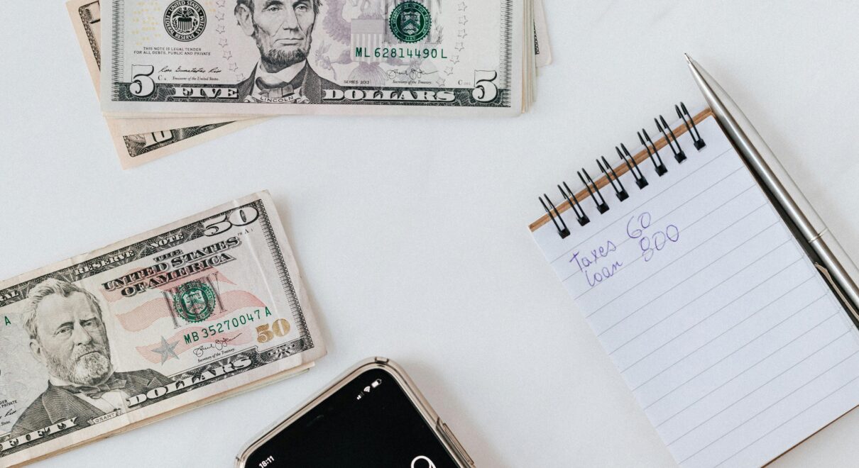 Overhead view of a hand using a calculator with US dollars and notes, representing finance and budgeting.