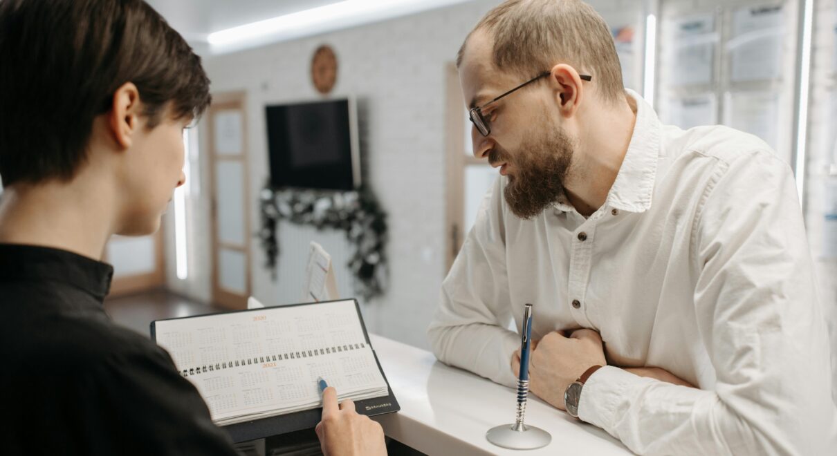 A receptionist and client converse over an appointment book at a clinic reception desk.